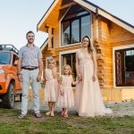 Family of four happily standing outside their modern wooden house on a sunny day.